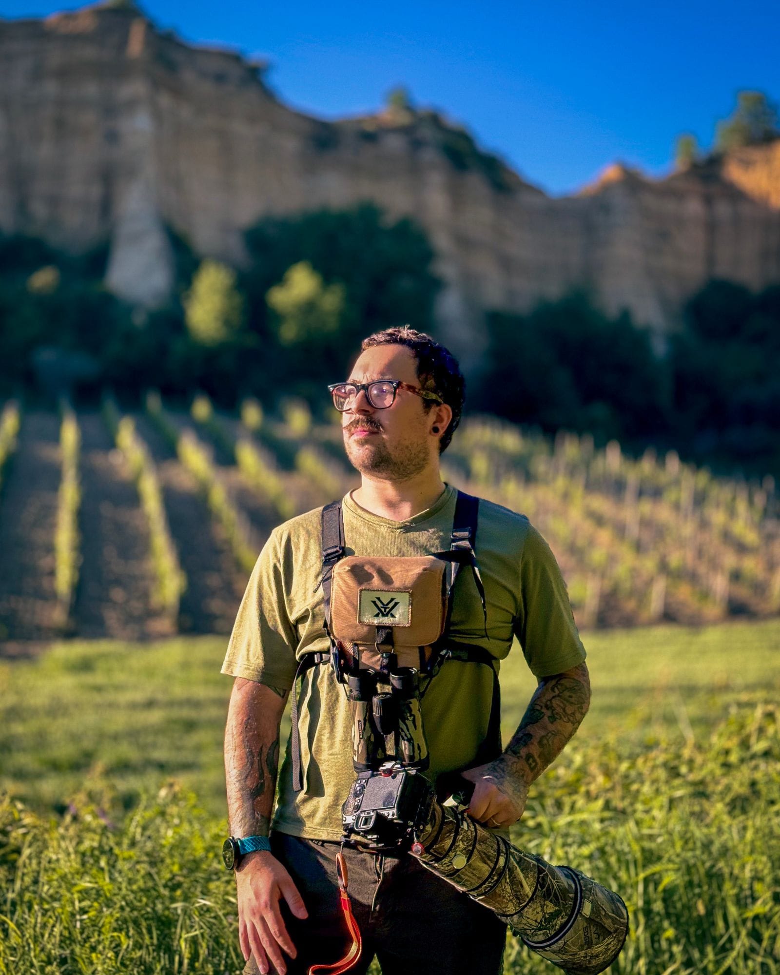 Giulio Fabbri sul campo con binocolo e teleobiettivo mimetico, davanti a una parete di roccia e a un vigneto